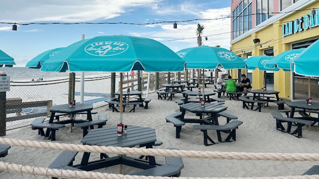 Turquoise branded commercial patio umbrellas shading black picnic tables at a beachfront restaurant with outdoor dining and ocean views.